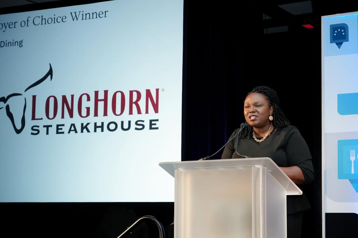 A woman speaks at a podium with a presentation slide behind her. The slide shows "Employer of Choice Winner" and the LongHorn Steakhouse logo.